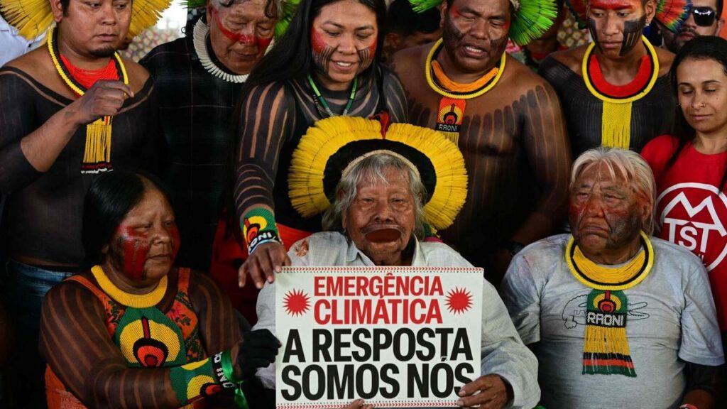 TOPSHOT - Brazilian indigenous leader and environmentalist Chief Raoni holds a sign reading "Climate emergency. We are the answer" during a press conference within a protest boat trip in the sidelines of the COP30 UN Climate Change Conference in Belem, Para State, Brazil, on November 12, 2025. Over 150 boats with members of indigenous communities and social organizations will sail the Guama River as part of a symbolic crossing during the COP30, according to organizers. (Photo by Pablo PORCIUNCULA / AFP)
