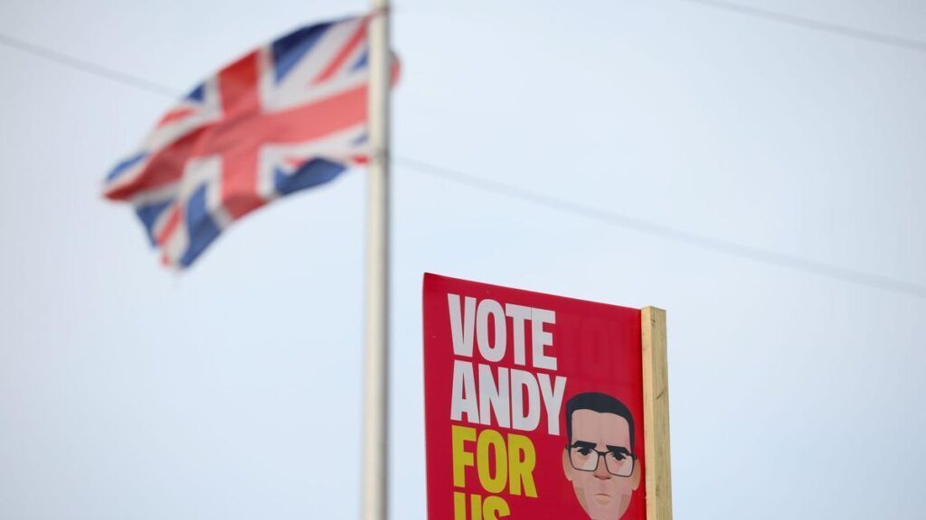 Una bandera del Reino Unido ondea cerca de un cartel del candidato laborista Andy Burnham en Wigan, Reino Unido, en una imagen del 15 de junio de 2026. La circunscripción inglesa de Makerfield celebra este jueves una elección parcial para elegir a su nuevo diputado, cita en la que Burnham concurre como candidato laborista a fin de entrar en el Parlamento y desafiar eventualmente el liderazgo de Keir Starmer. EFE/EPA/ADAM VAUGHAN